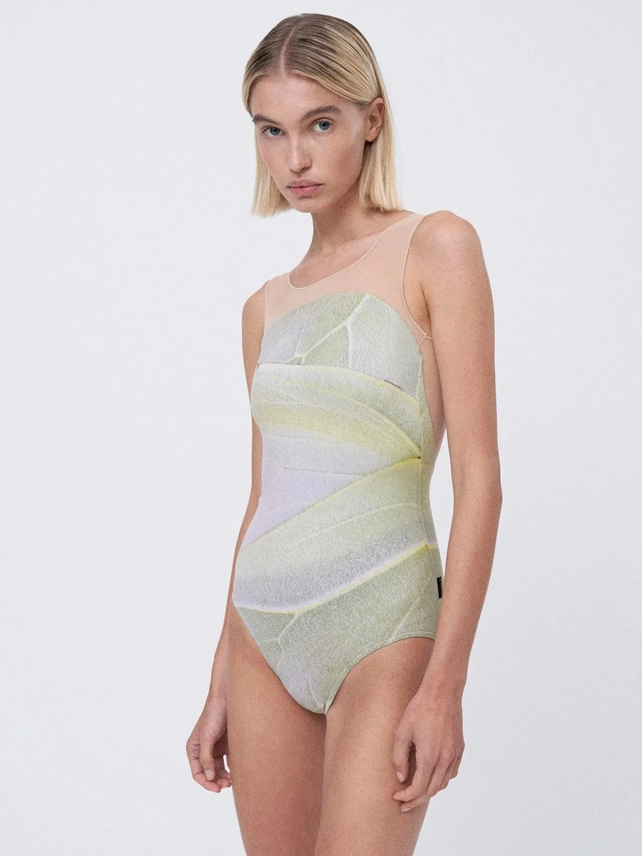 A young woman with blonde hair in a bob haircut wearing a sleeveless body suit with a pastel leaf pattern, standing against a plain white background.