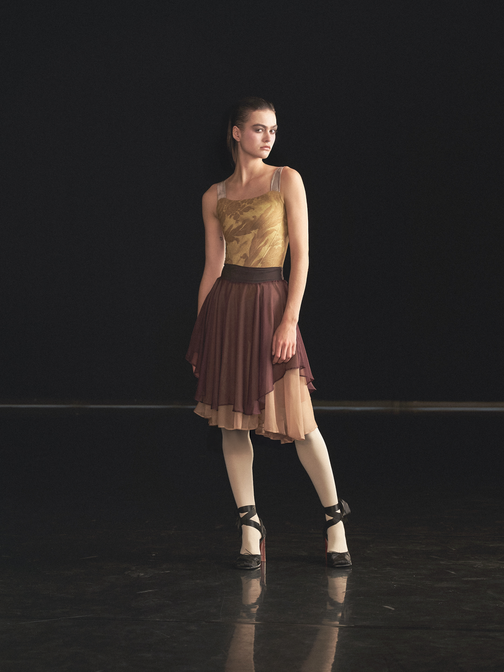 Maldire Alta Corsetteria campaign: dancer in a golden corset leotard and layered skirt on a dark stage