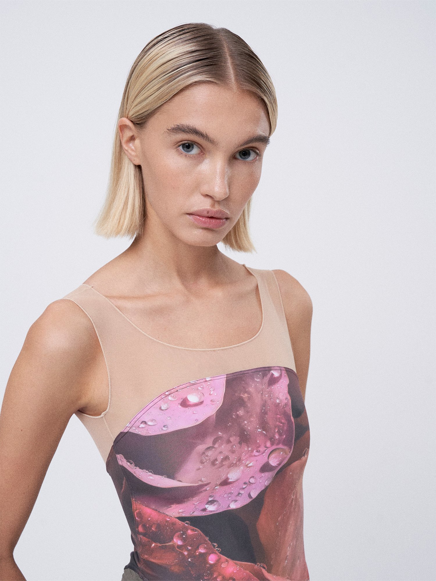 A young woman with short blonde hair and blue eyes looking directly at the camera. She is wearing a sleeveless top with a graphic print featuring pink and purple tones and waterdrop effects, set against a neutral background.