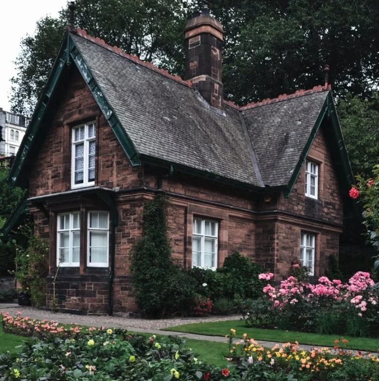 A small, historic brick house with a steeply pitched roof and white-framed windows, surrounded by a well-kept garden with colorful flowers and lush greenery.