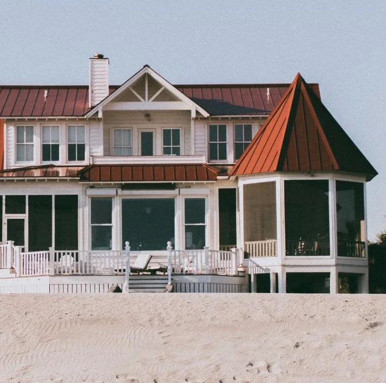 Large beachfront house with white exterior, multiple windows, and a red metal roof, featuring a screened-in porch and a tower-like structure with a pointed roof, situated on sandy terrain.