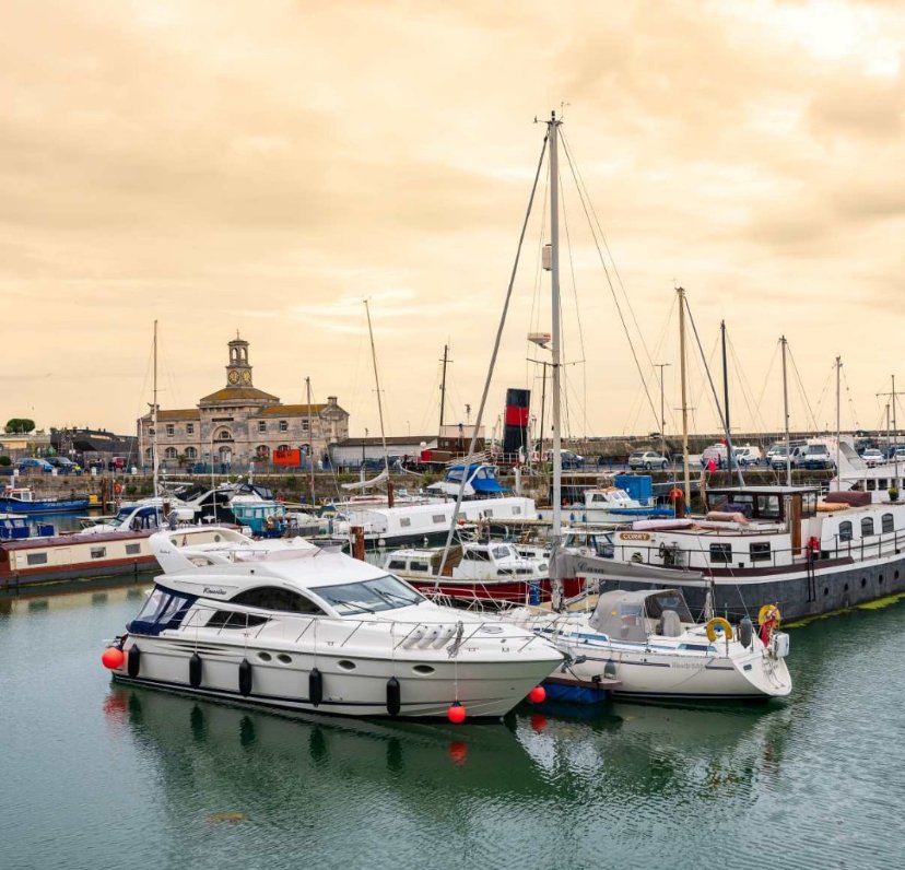 A marina with various boats and yachts docked, with a building and overcast sky in the background.