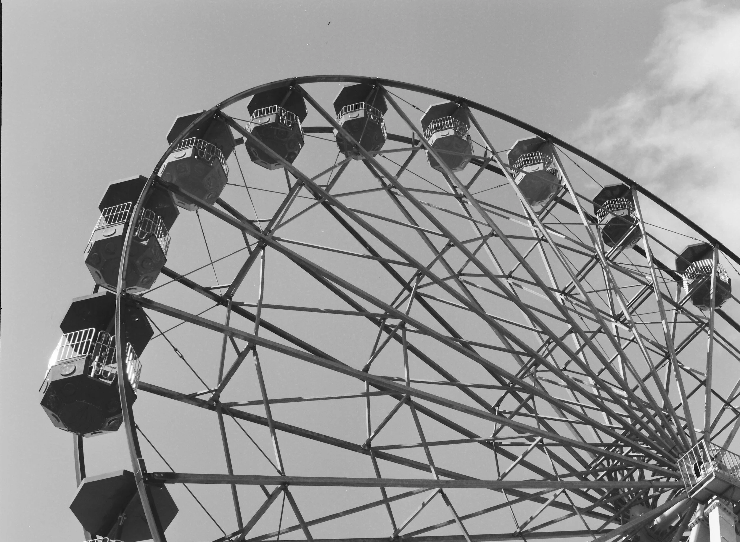 A black and white photograph of a large Ferris wheel with multiple enclosed gondolas against a partly cloudy sky.