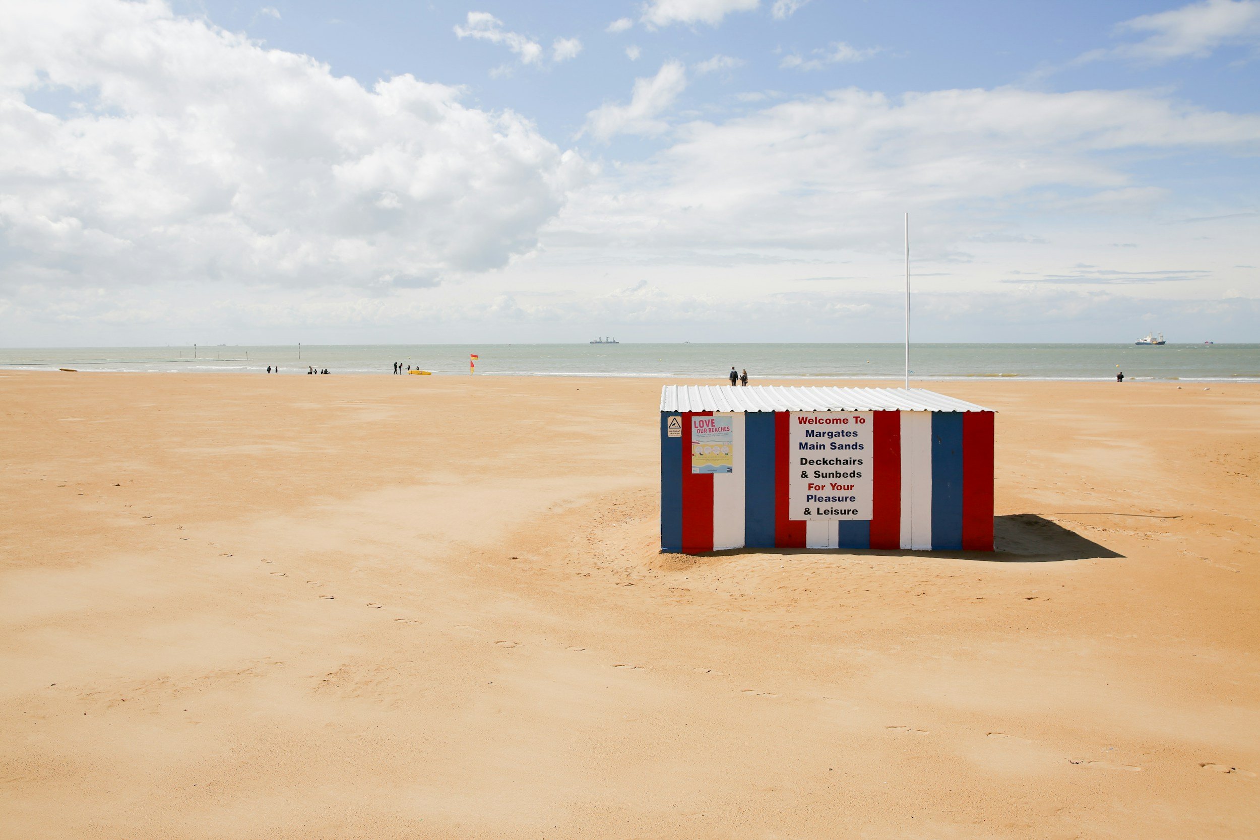 A sandy beach with a colorful striped beach hut and a sign welcoming visitors to Margate's main sands, deckchairs, and sunbeds for leisure, with the ocean and a cloudy sky in the background.