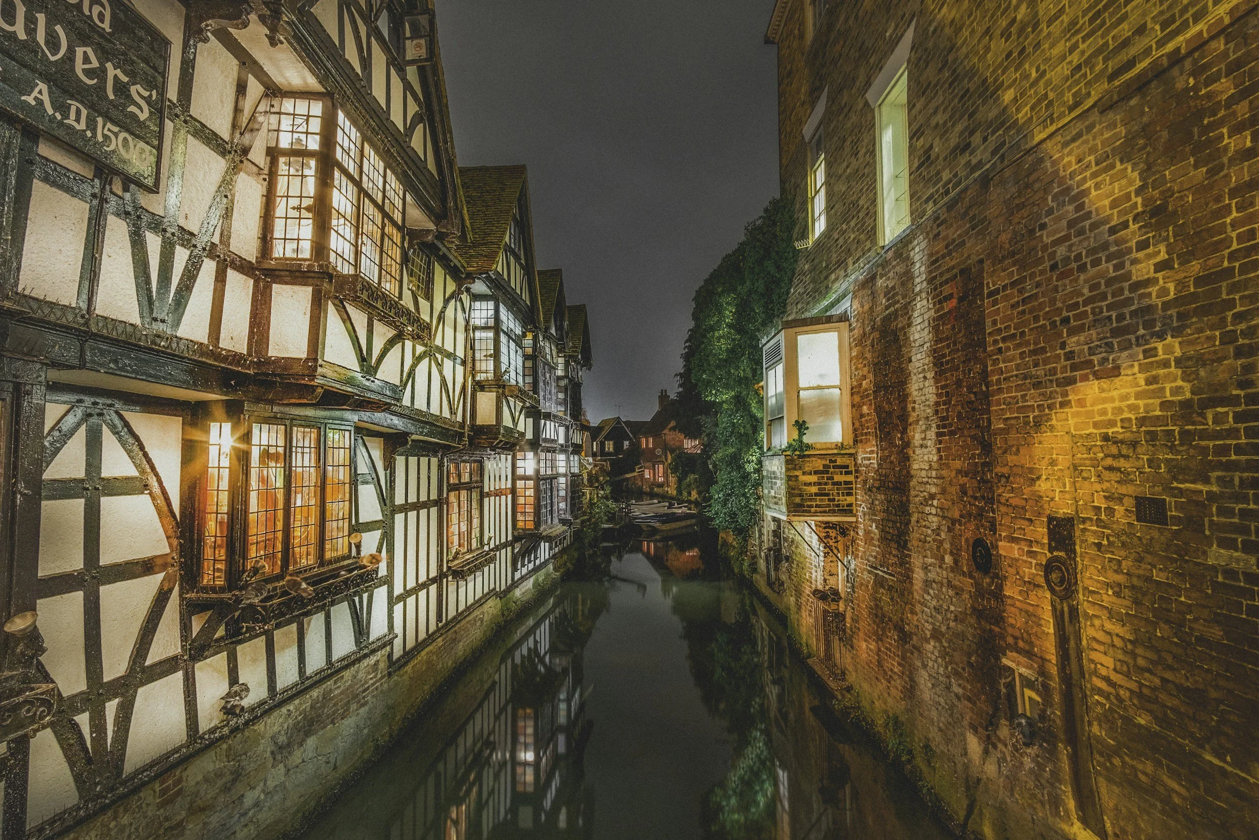 Nighttime scene of a narrow canal between historic Tudor-style buildings with illuminated windows on the left and brick buildings on the right, reflecting in the water.