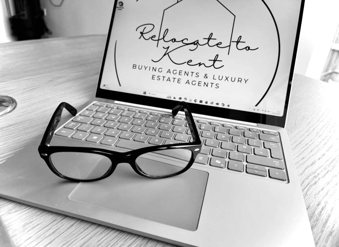 A laptop on a wooden desk with a pair of glasses resting on the trackpad and the screen displaying a logo for "Refocate to Kent" with the text "Buying Agents & Luxury Estate Agents".
