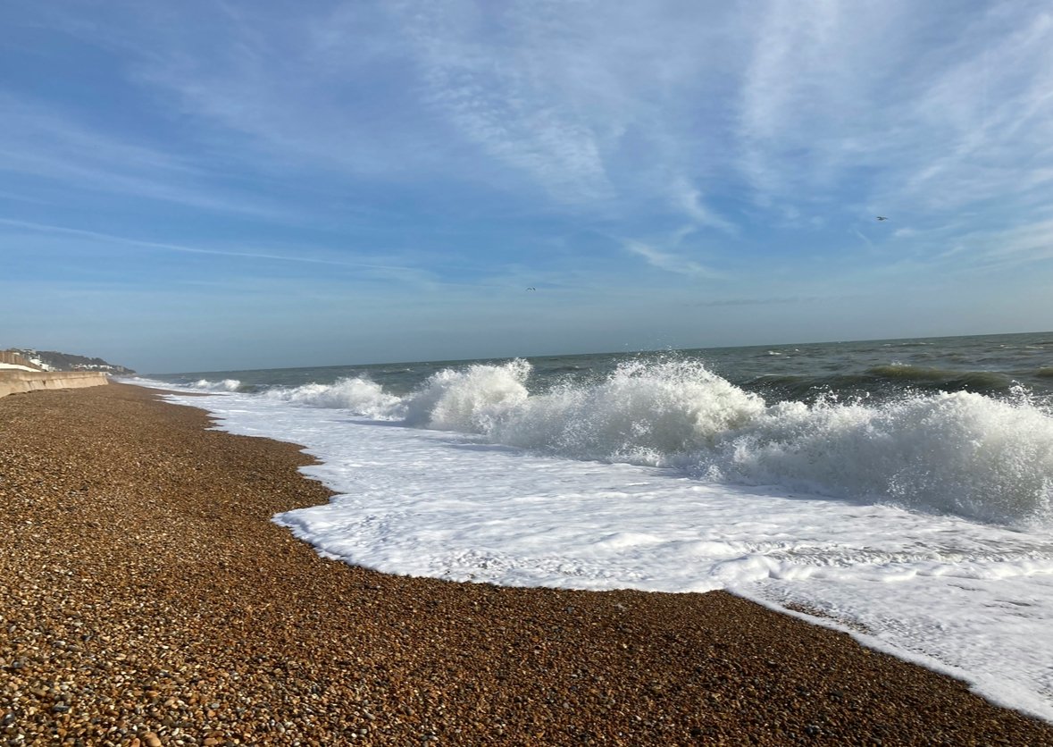 The image shows a pebble beach with waves crashing onto the shore under a partly cloudy sky, with some buildings visible in the distance on the left.