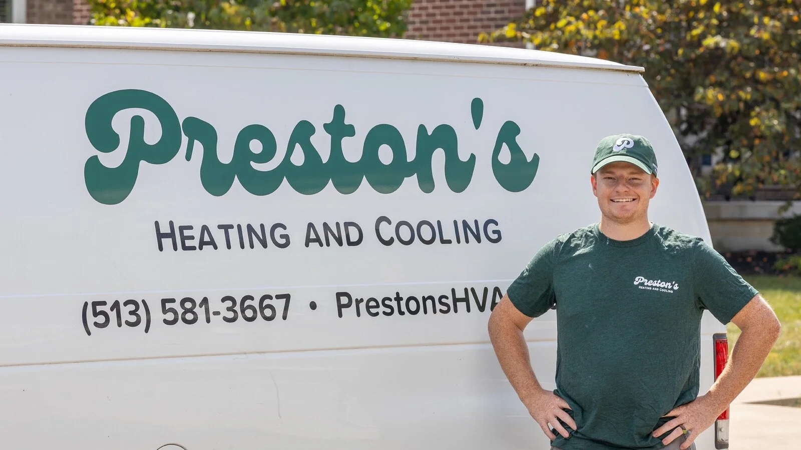 A smiling man in a green Preston's Heating and Cooling T-shirt and cap standing next to a white van with Preston's Heating and Cooling logo and contact information printed on it.