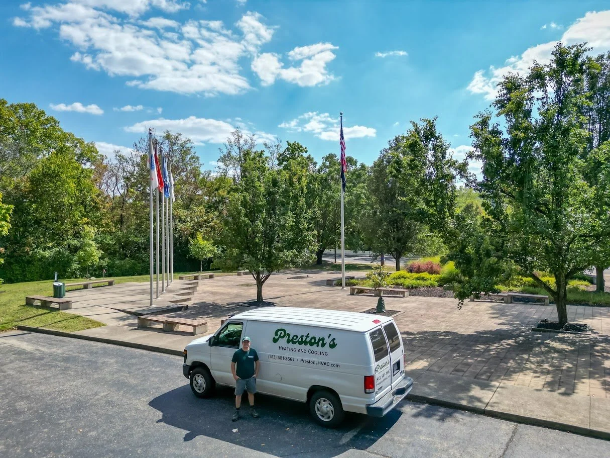 A man standing in front of a white van for Preston's Heating and Cooling. The van is parked in a lot near a park with trees, flagpoles, and benches, on a sunny day with a partly cloudy sky.
