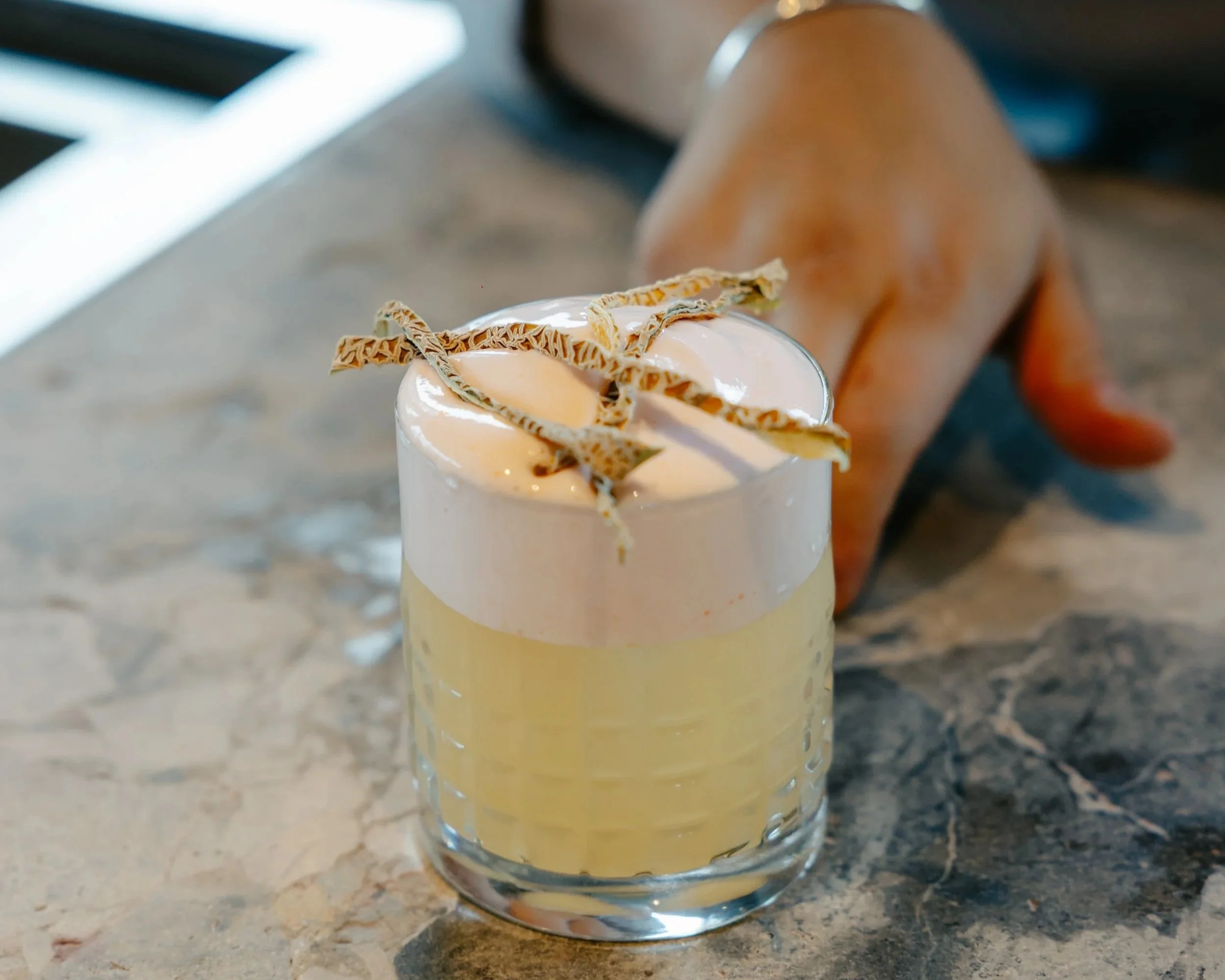A layered cocktail with a frothy top, garnished with chocolate curls in a clear glass on a marble surface.