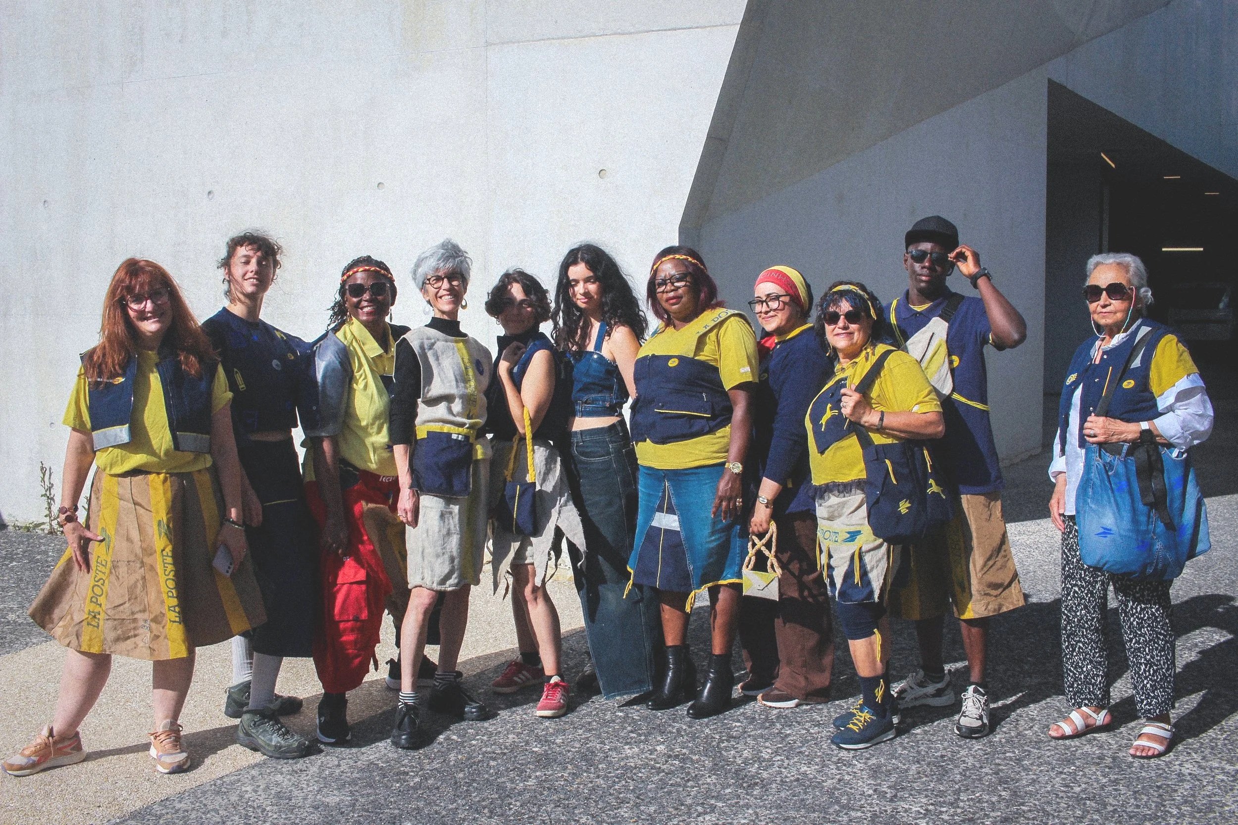Groupe de personnes posant pour une photo à l'extérieur, portant des vêtements jaunes et bleus, certains avec des lunettes de soleil, devant un bâtiment en béton.