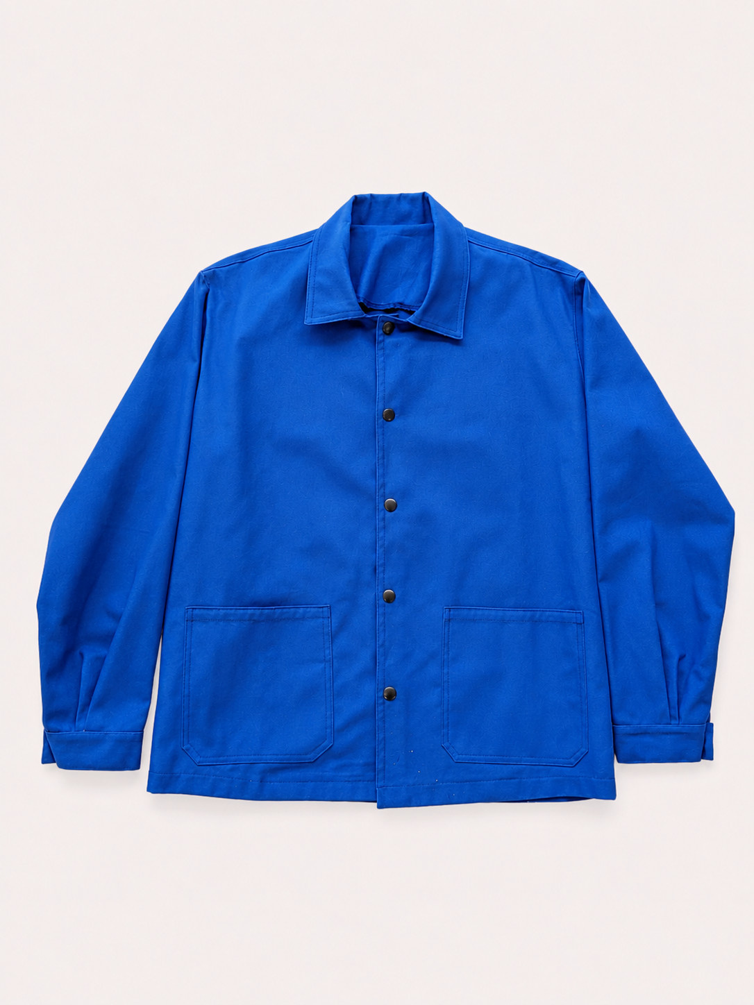 Veste bleue en coton à manches longues avec poches frontales, boutons et col classique, suspendue contre un fond blanc
