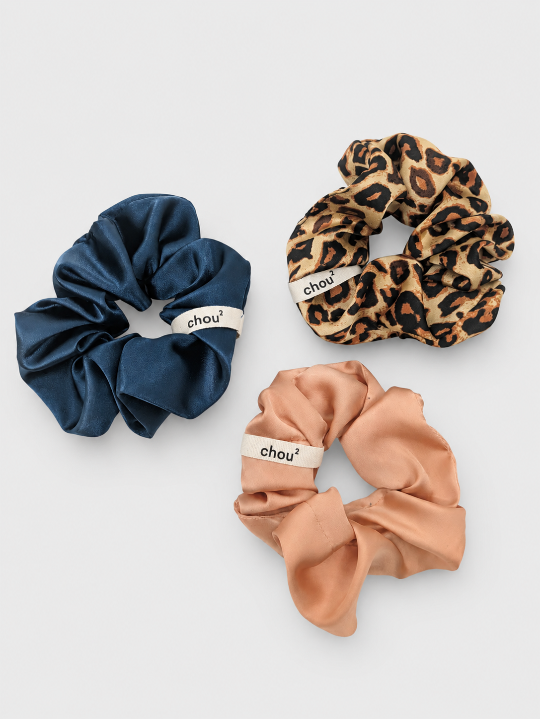 Trois scrunchies en satin de différentes couleurs et motifs, chacun avec une étiquette marquée 'chou²'.