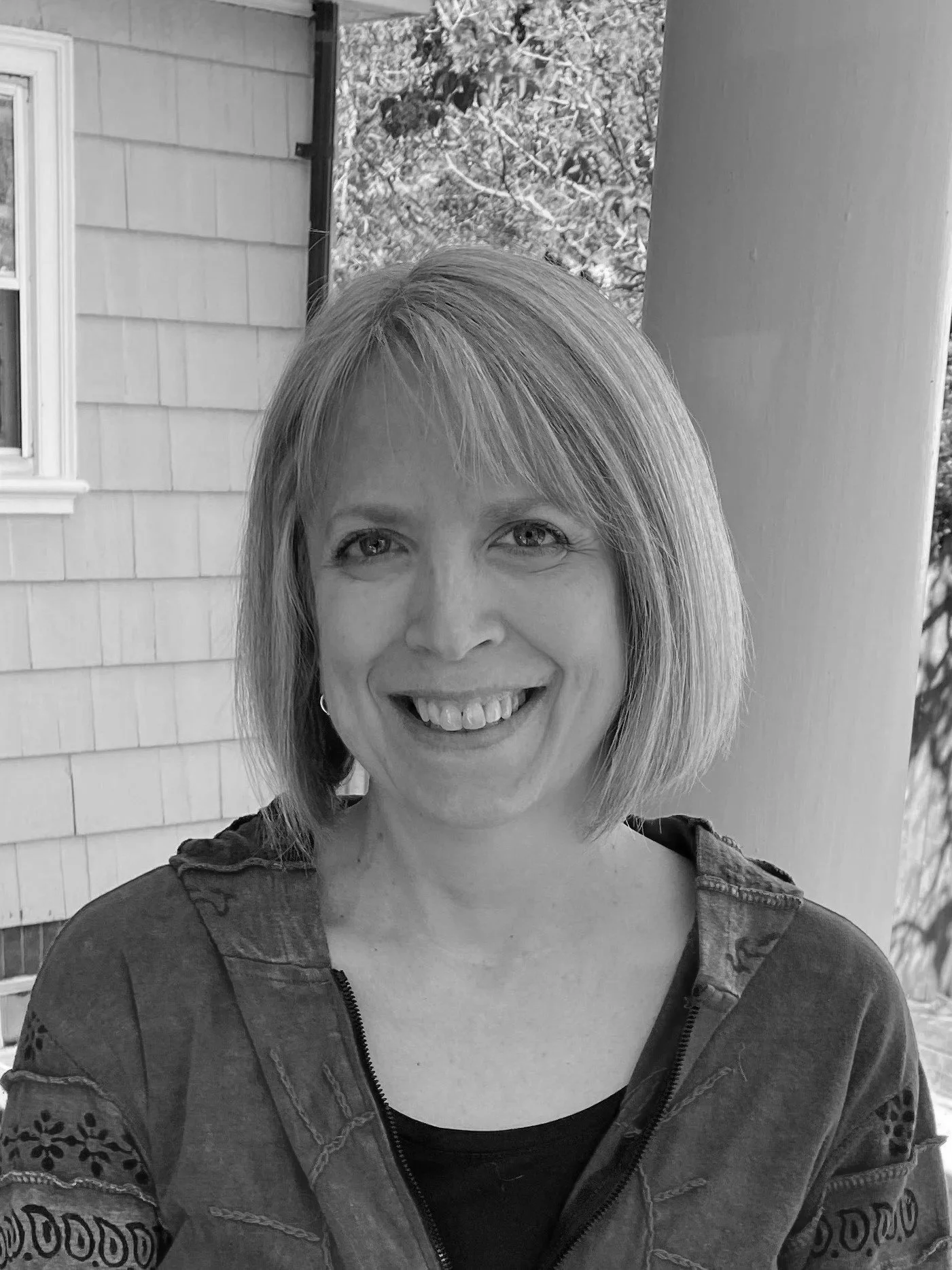 A smiling woman with short light-colored hair and light eyes, wearing a jacket with embroidered details, standing outside in front of a porch and trees, in black and white.