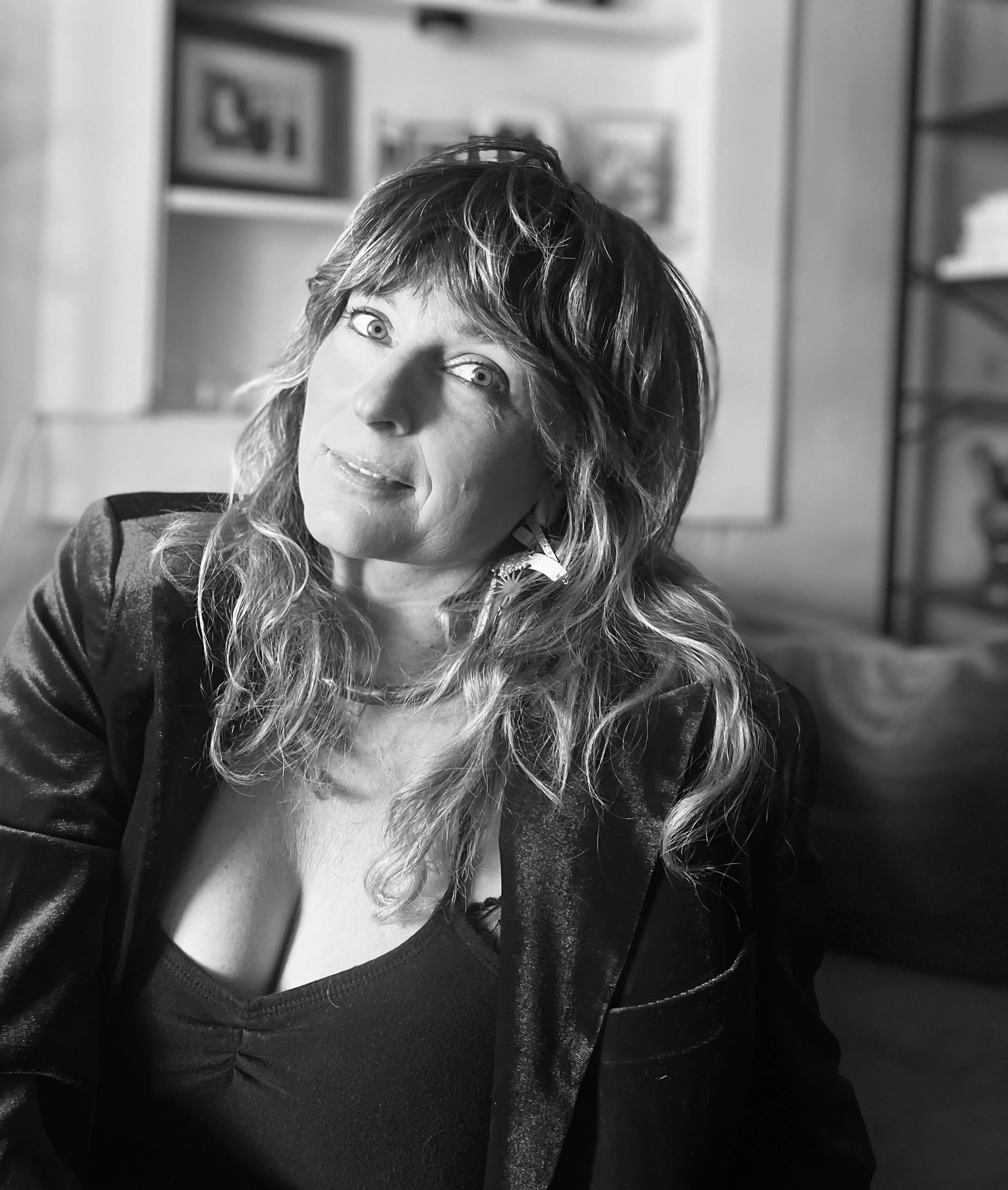 Black and white portrait of a woman with wavy hair, wearing a dark, textured jacket and a necklace, sitting in an indoor space with blurred shelves or picture frames in the background.
