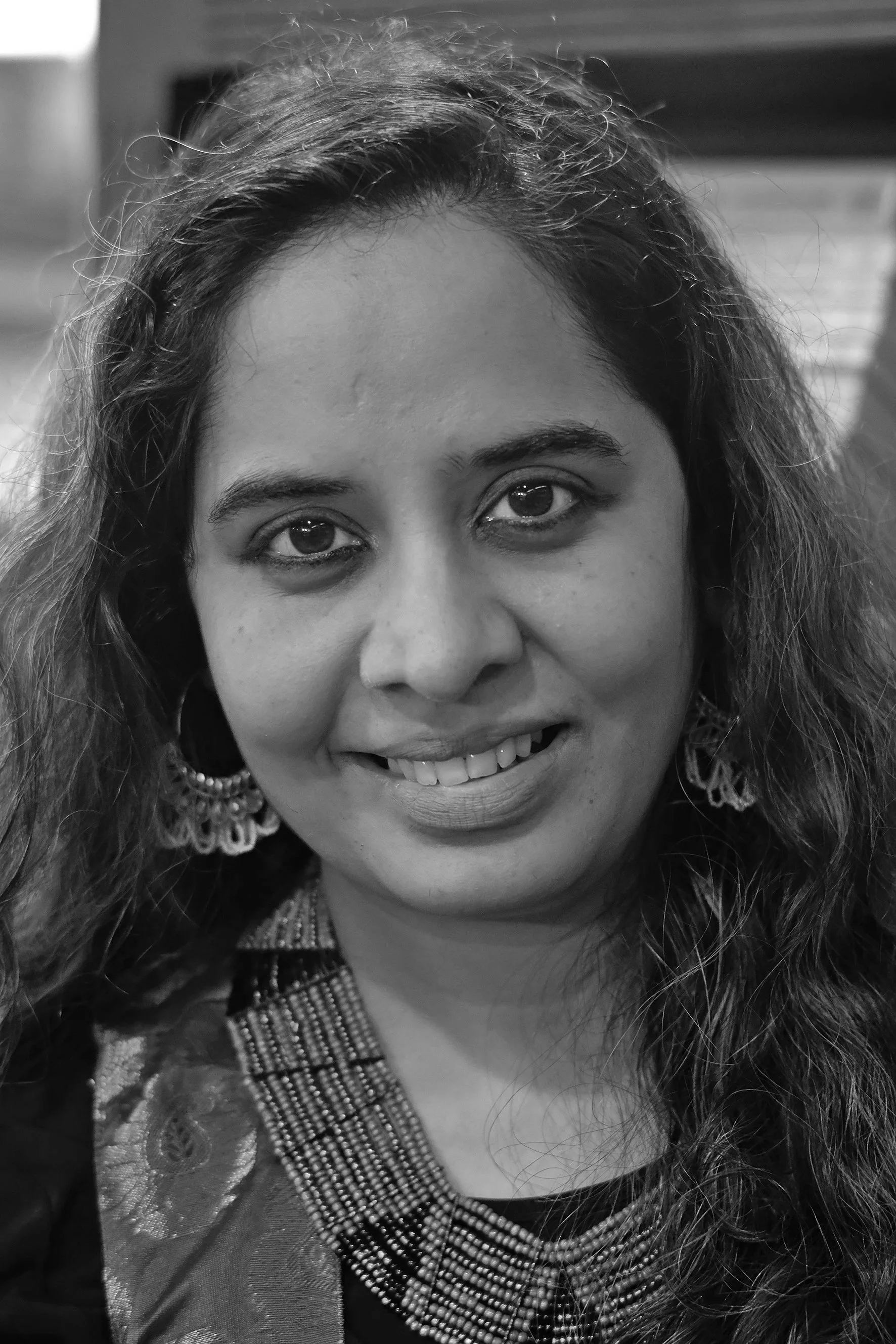 Close-up black and white photo of a woman smiling, wearing earrings and traditional attire with intricate patterns.