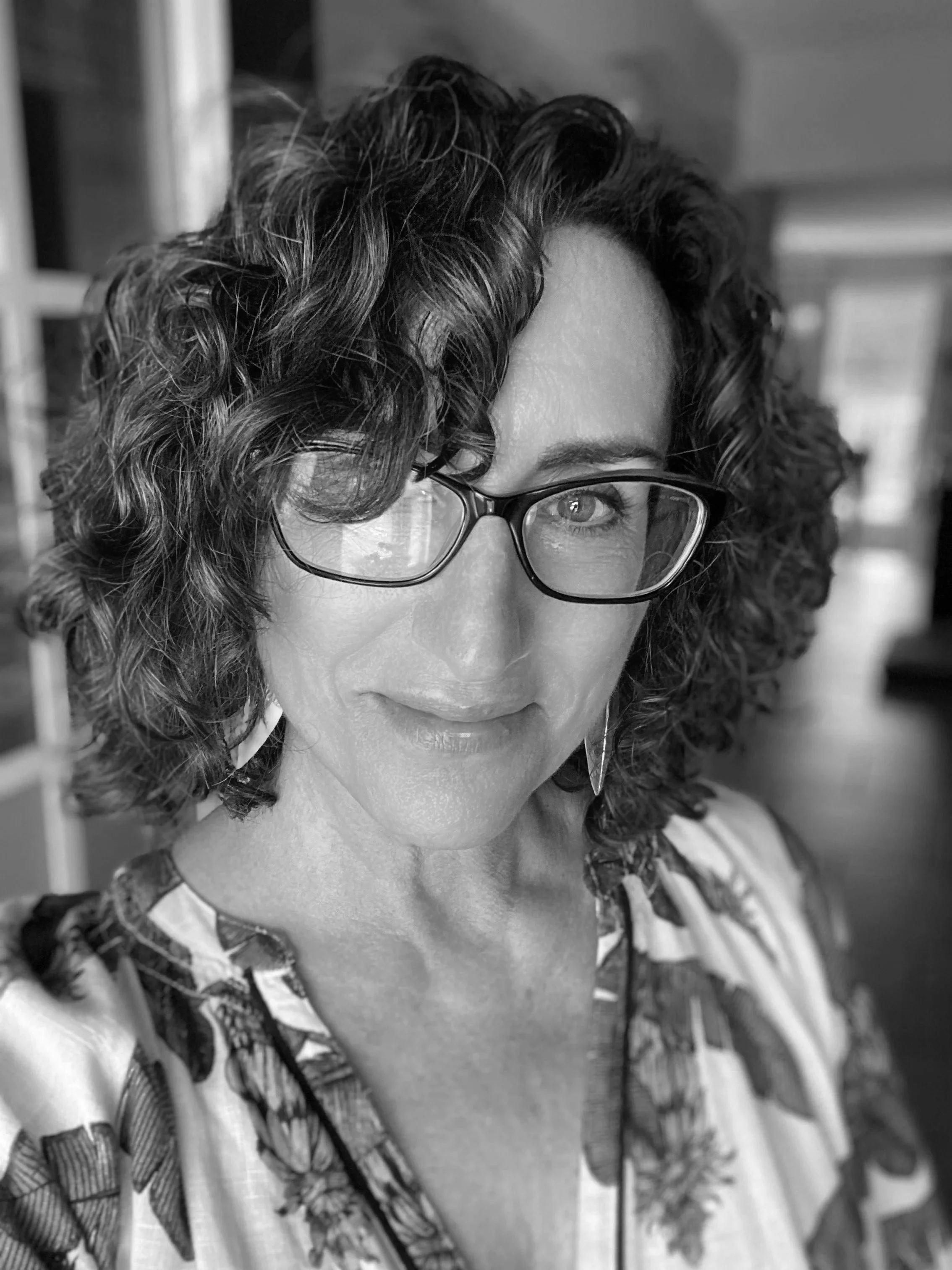 A woman with curly hair and glasses, looking at the camera in a black-and-white photo.