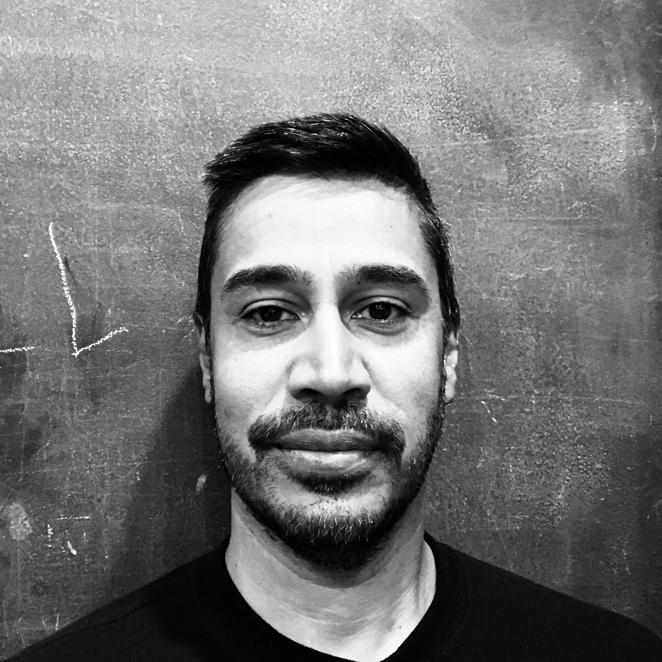 Close-up black and white photo of a part of a man's face with dark hair, eyebrows, eyes, nose, and beard, against a textured wall background.