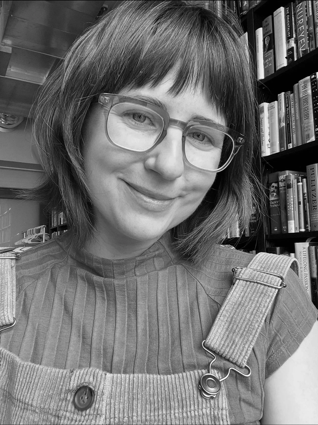 A woman with short hair and glasses smiling in a library surrounded by books.