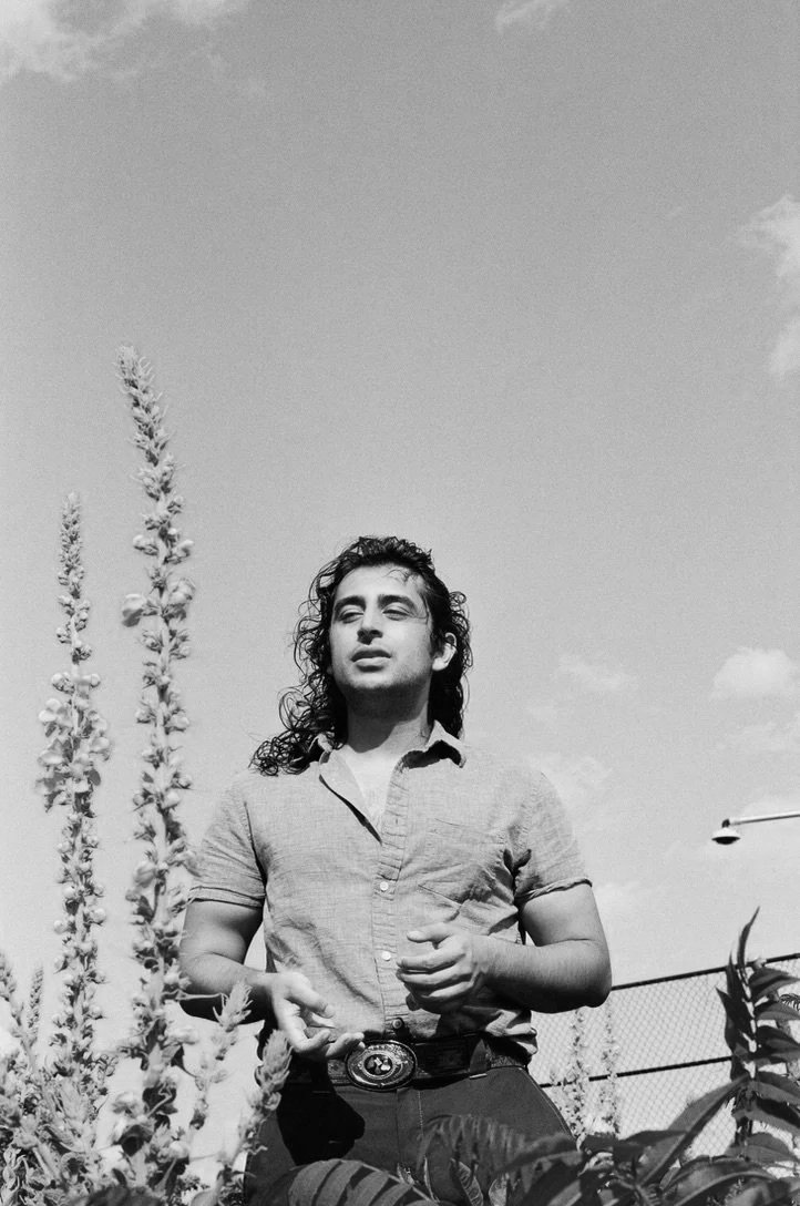 A person with curly hair standing outdoors near tall plants, wearing a short-sleeved button-up shirt with a camera hanging around their neck.