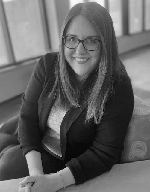 A woman with long hair and glasses smiling, sitting indoors on a cushioned chair.