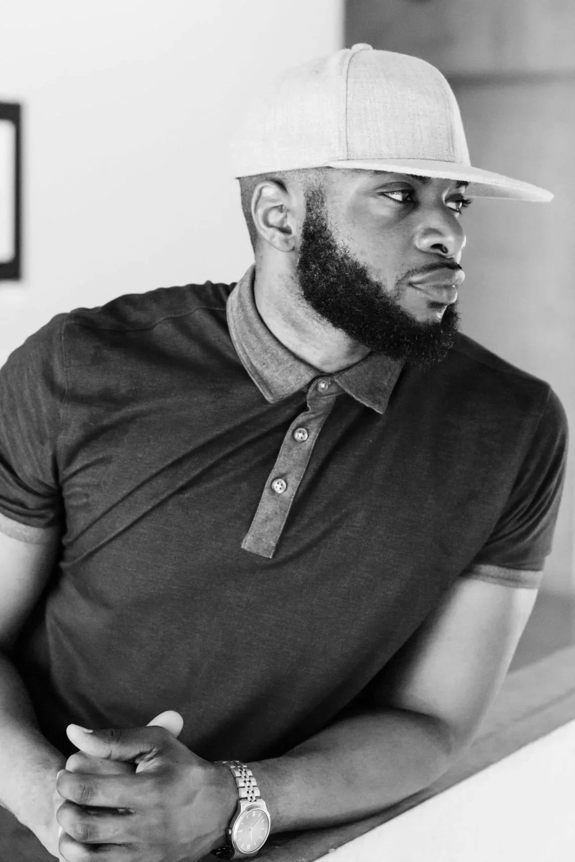 A man with a beard wearing a light-colored baseball cap, a dark polo shirt with a contrasting collar, and a wristwatch, looking to the side.
