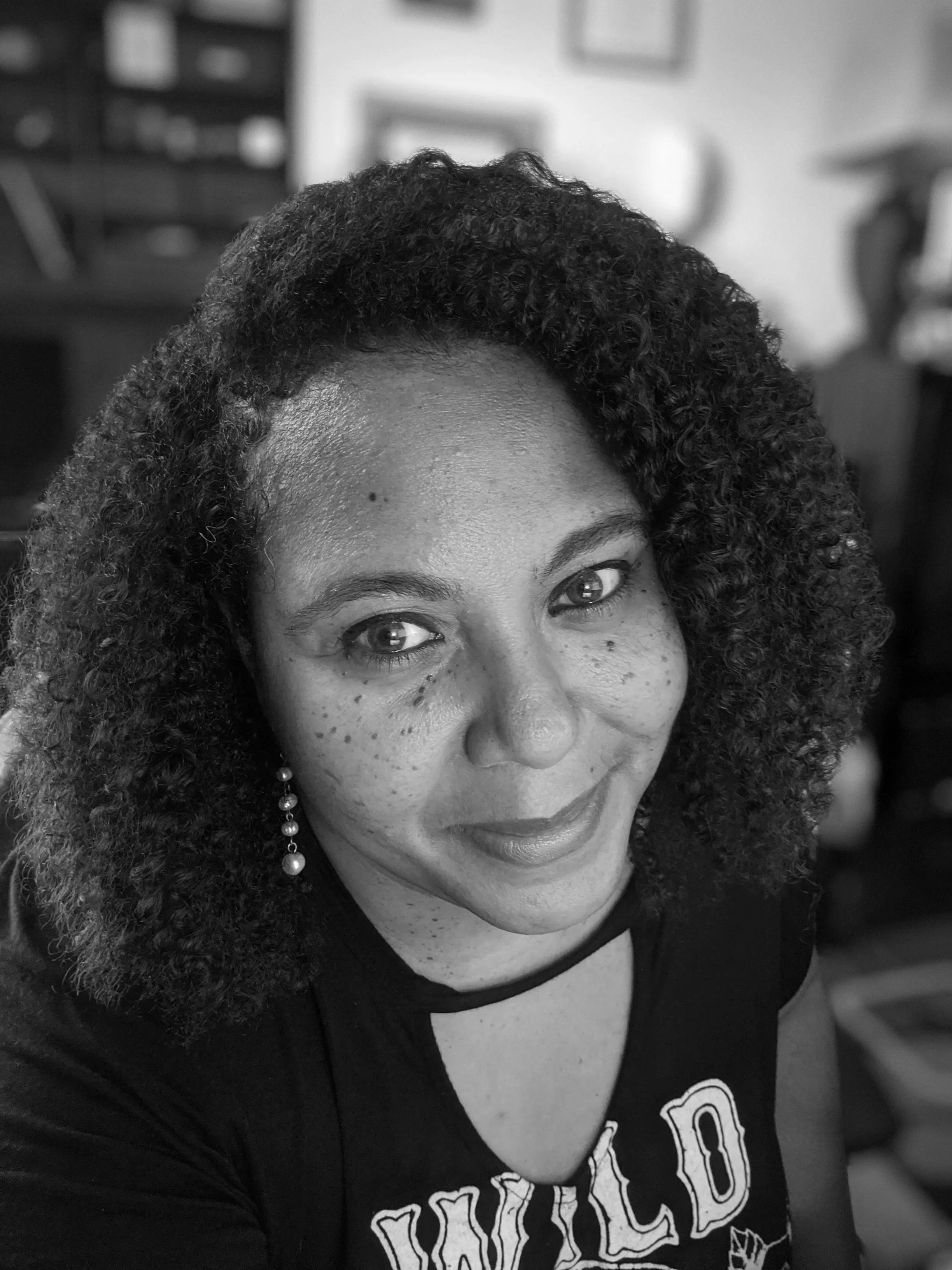 A black and white close-up photo of a woman with curly hair, smiling, wearing earrings, and a graphic T-shirt, in an indoor setting.