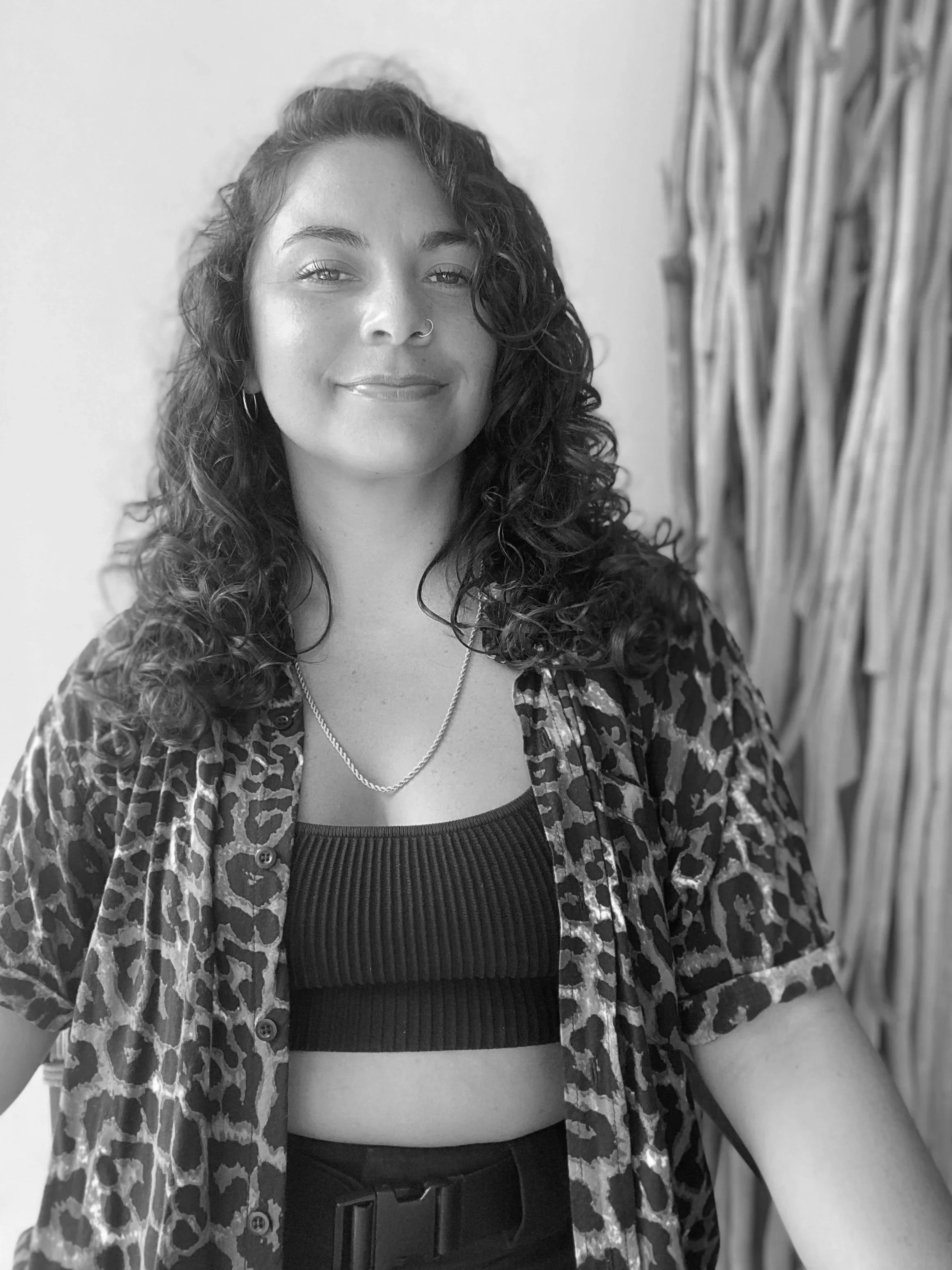 A woman with curly hair, wearing a patterned animal print shirt over a ribbed tank top, accessorized with a necklace, hoop earrings, and a nose ring, standing in front of a plain wall and a bundle of tall plants.