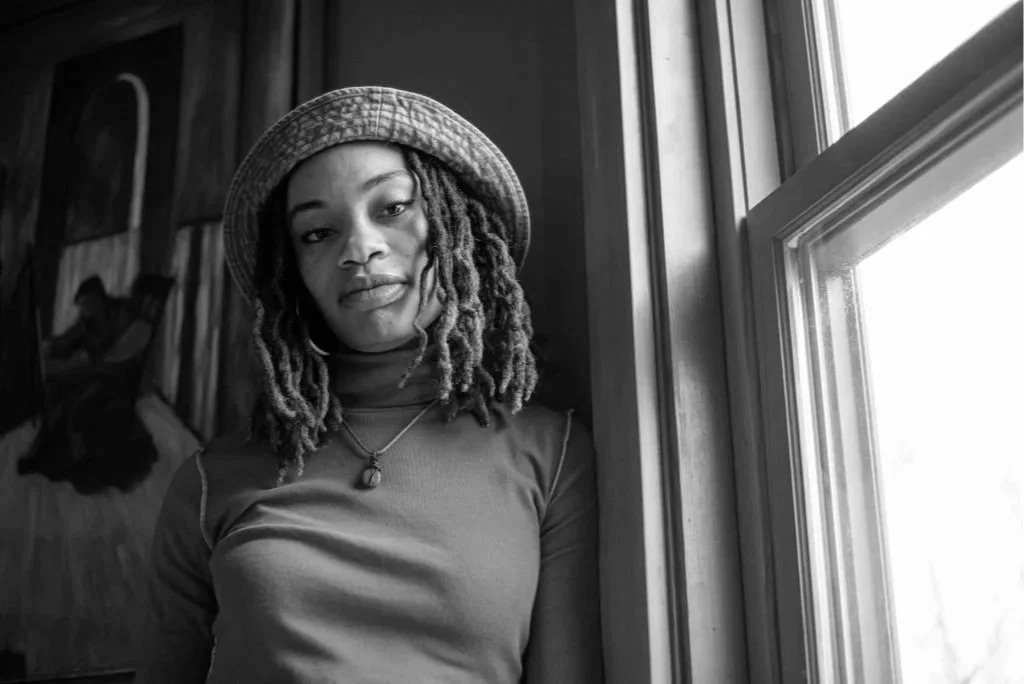 A young woman with shoulder-length dreadlocks, wearing a hat, standing indoors by a window, in a black-and-white photo.
