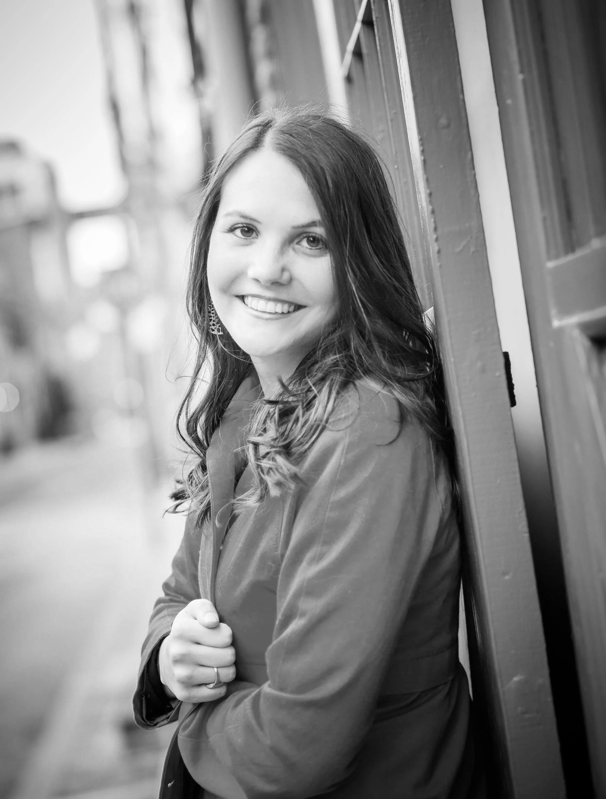 A young woman with long hair smiling, leaning against a wooden wall, wearing a jacket, with one hand resting on her jacket. The photo is in black and white.