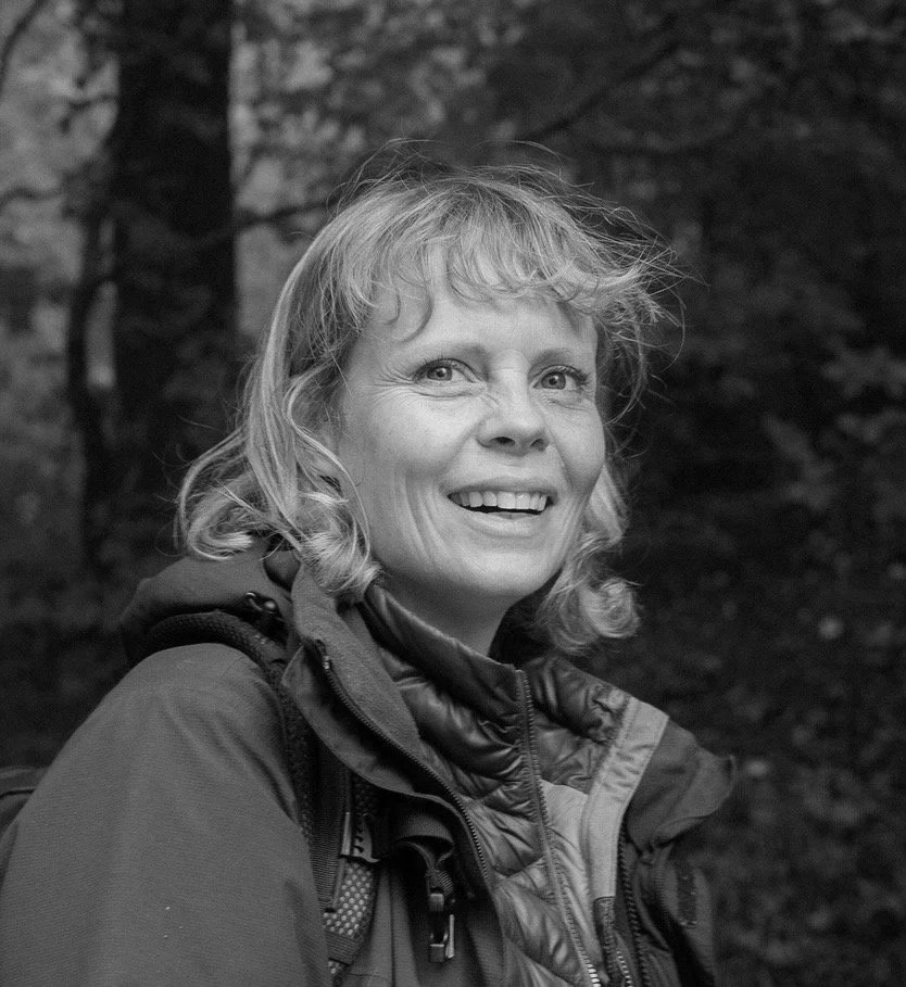 A smiling middle-aged woman with wavy, shoulder-length hair, wearing outdoor gear, in a forest setting.