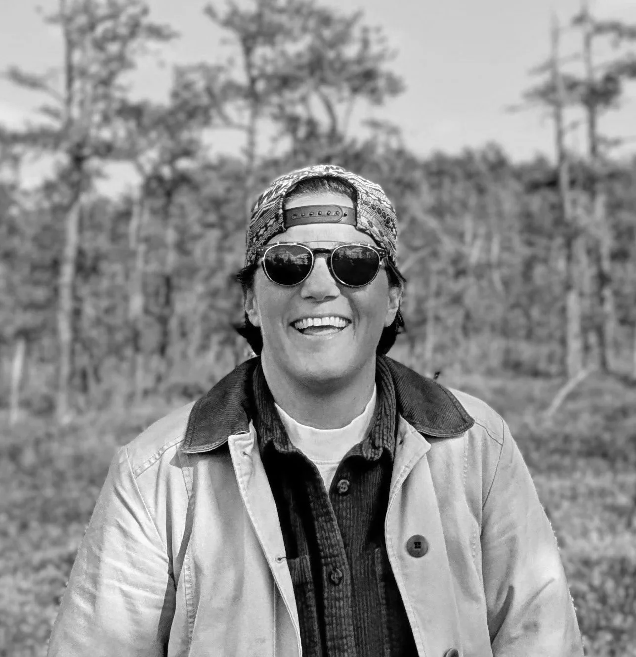 A man smiling outdoors wearing sunglasses, a patterned cap backward, a light-colored jacket, and a collared shirt, with trees in the background.