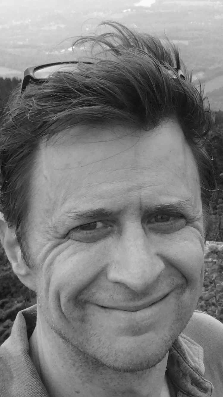 Close-up of a smiling man with short, wavy hair, wearing sunglasses on his head, outdoors in a rural or mountainous area.