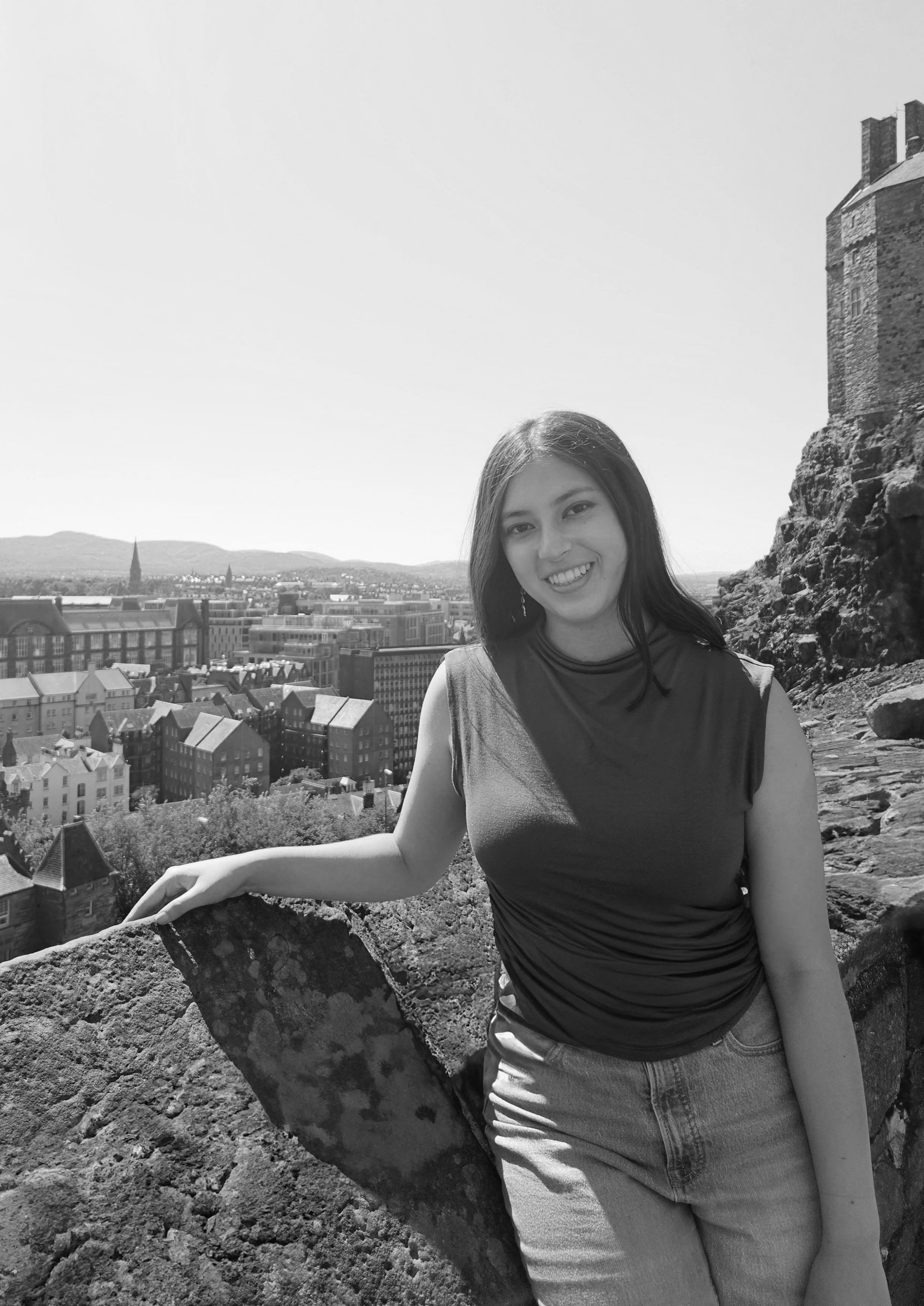 A young woman smiling outdoors on a sunny day, with a cityscape and mountains in the background, standing next to a large stone structure.