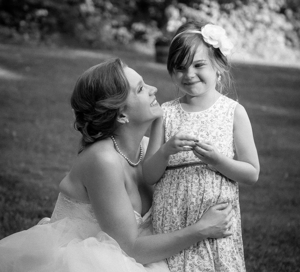A woman and a young girl smiling at each other outdoors, the woman is kneeling and the girl is standing, both dressed in light-colored dresses, with the girl wearing a large flower hair accessory.