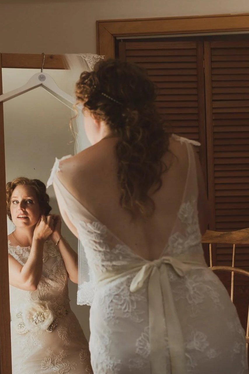 A woman in a wedding dress looking at herself in a mirror, adjusting her earrings.