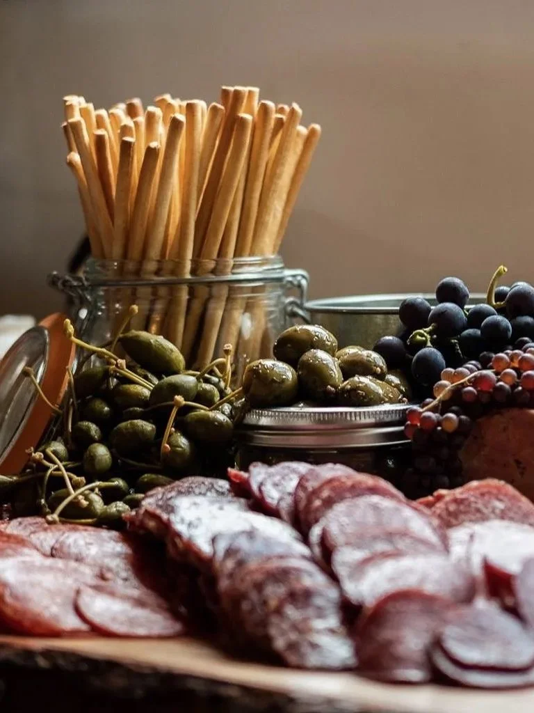 Charcuterie board with sliced meats, grapes, and olives, with breadsticks in a jar in the background.