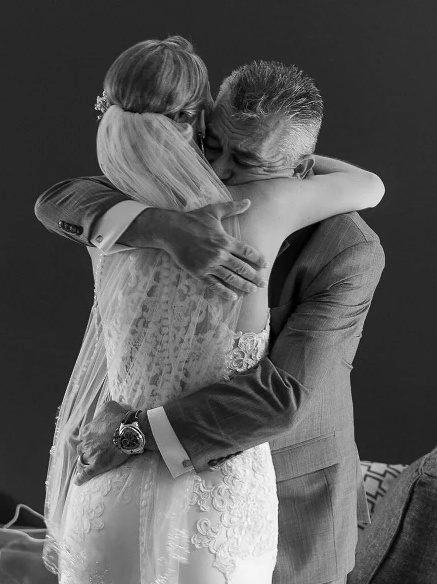 A woman dressed in a wedding gown and a man in a suit embrace and hug each other. The woman wears a lace dress and a veil, and the man is wearing a suit jacket, dress shirt, and a watch. They are sharing an emotional moment.