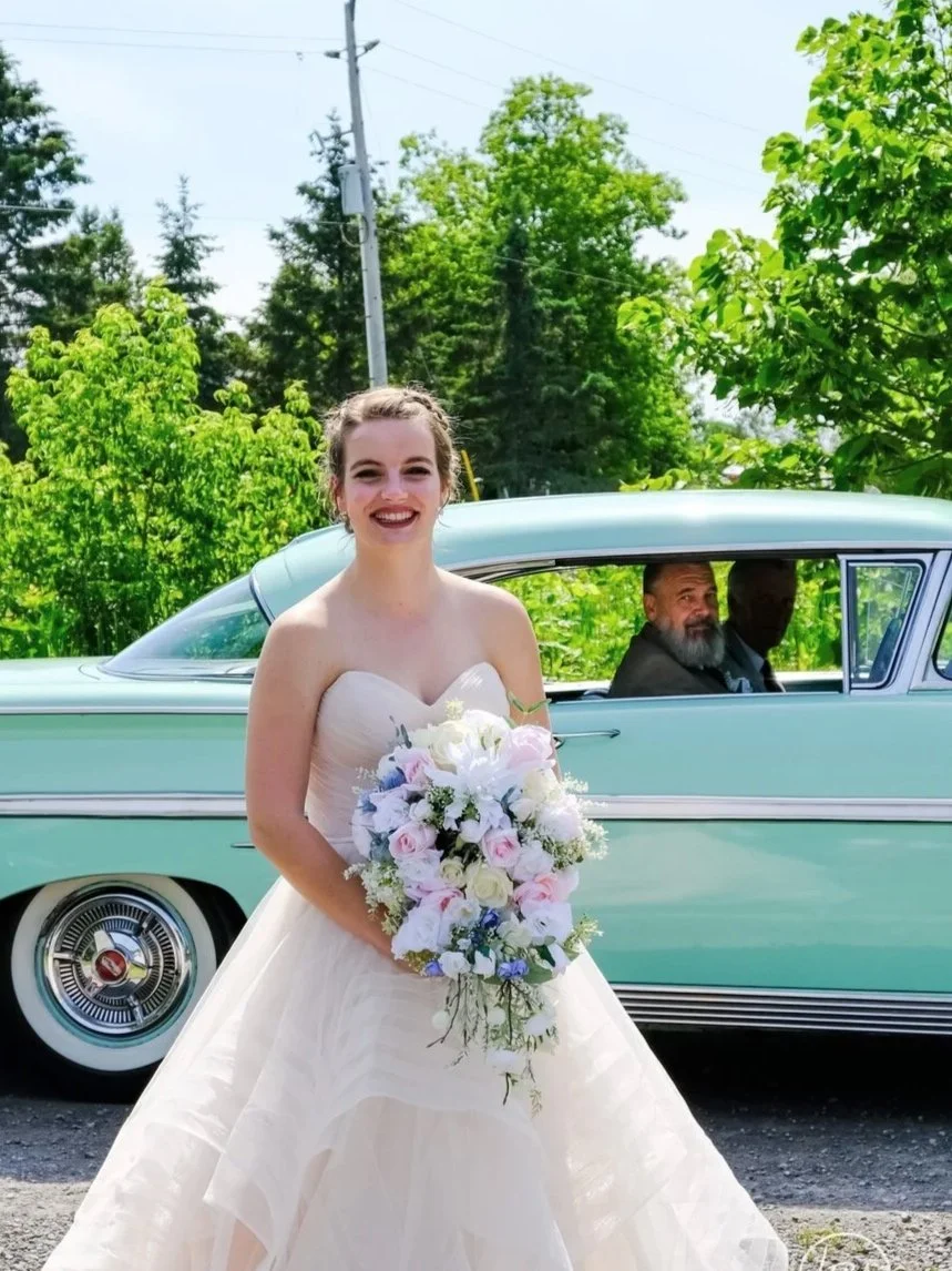 A bride in a white wedding dress holding a bouquet of white and pink flowers, smiling outdoors next to a vintage light blue car with two men inside, surrounded by green trees and utility poles.