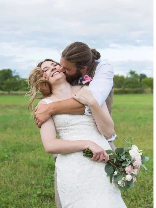 A happy couple, the bride with wavy blonde hair in a white wedding dress holding a bouquet, and the groom with long hair and a beard in a vest, embracing and kissing outdoors in a grassy field with a cloudy sky.