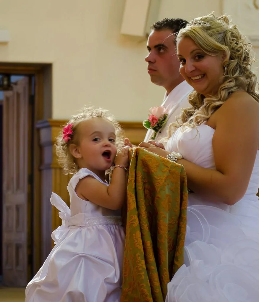 A bride with blonde curly hair smiling and wearing a white dress, holding a cloth, standing in a room with wooden paneling, beside a man with dark hair and a serious expression, and a young girl with curly blonde hair, wearing a white dress with a pi