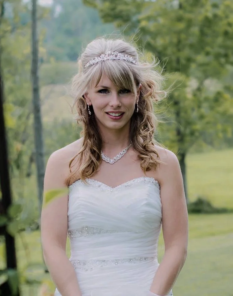 A bride in a white wedding dress standing outdoors with trees in the background, wearing jewelry and a tiara.