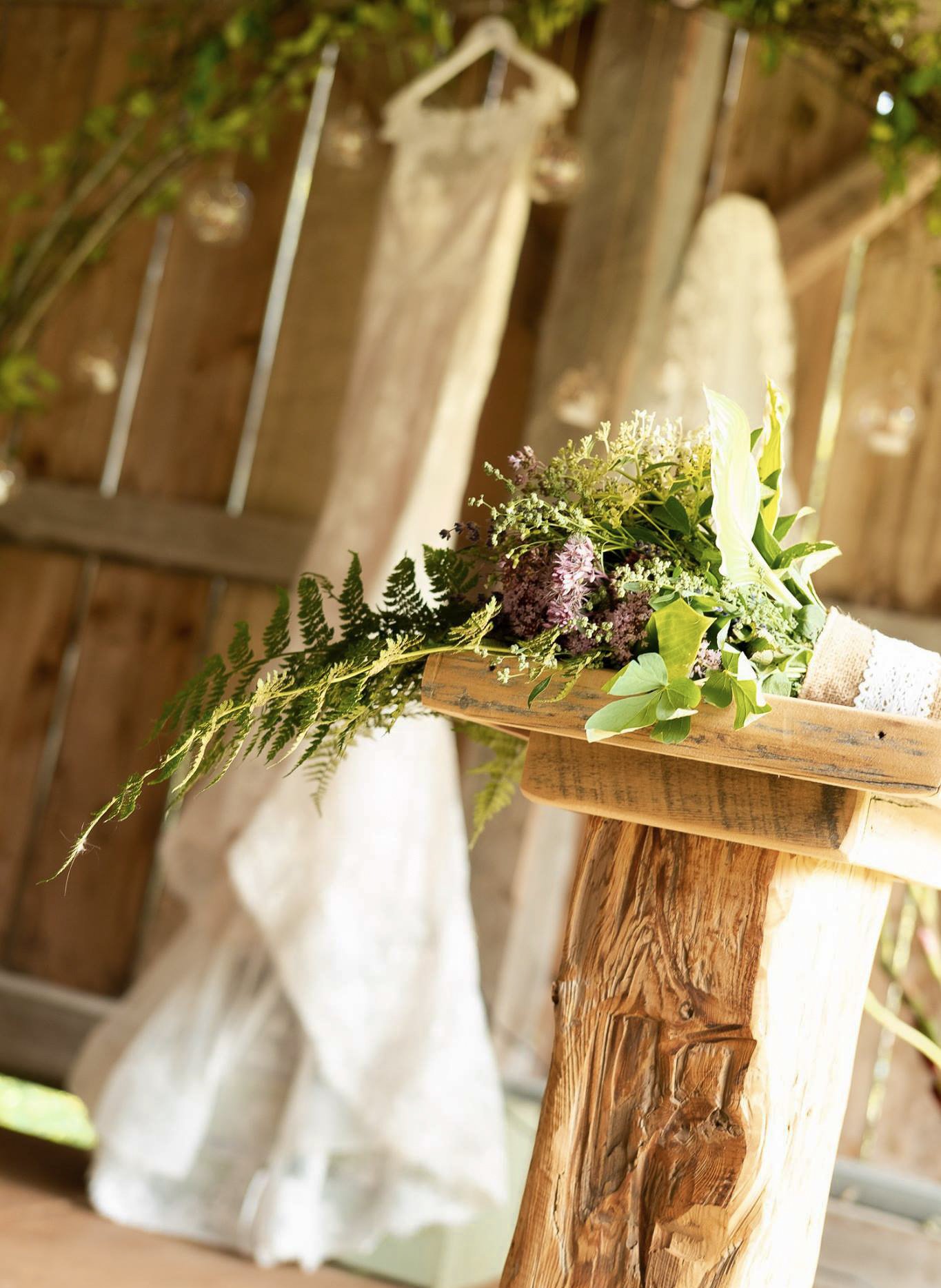 A small wooden shelf holding a flower arrangement with various greenery and flowers. The shelf is mounted on a rustic wooden wall with hanging ornaments in the background.