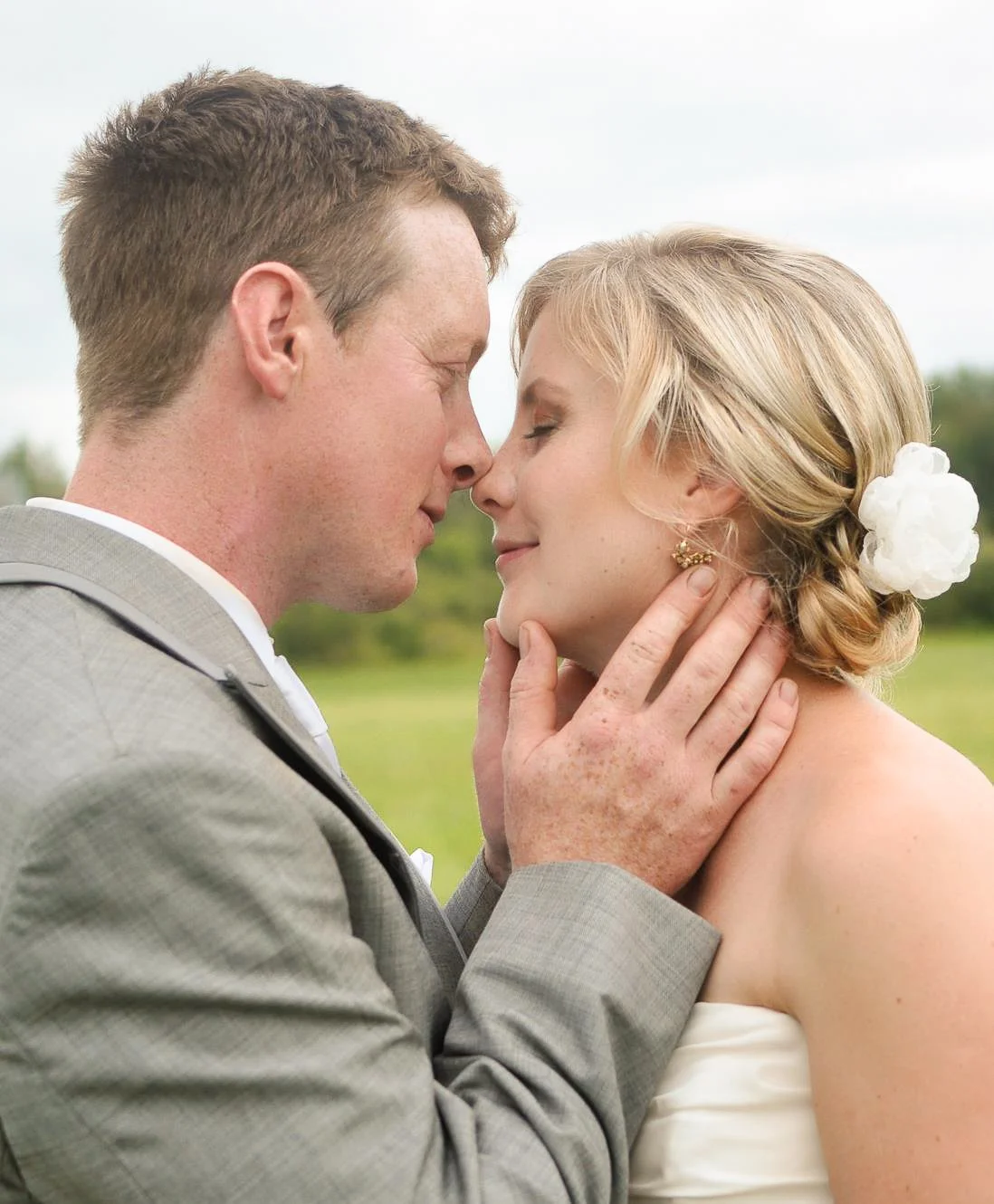 A bride and groom with eyes closed, gently touching foreheads and smiling during their wedding day outdoors.