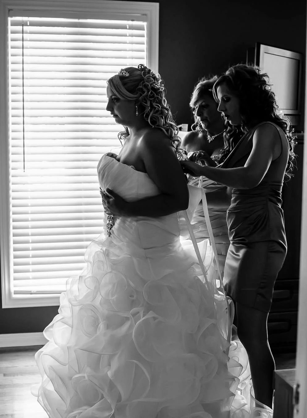A bride in a wedding gown is being assisted by two women, one helping with her dress and the other adjusting her hair, in a room with closed window blinds.
