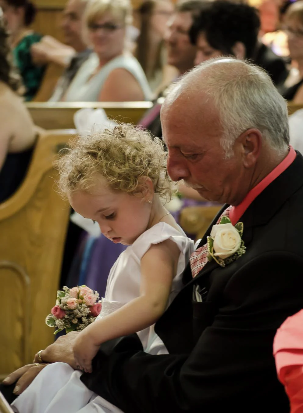 An elderly man with a white mustache and bald head in a black suit, pink shirt, and a boutonniere sitting in a church pew with a young girl with curly blonde hair in a white dress, holding a small bouquet of flowers, as they both look down.