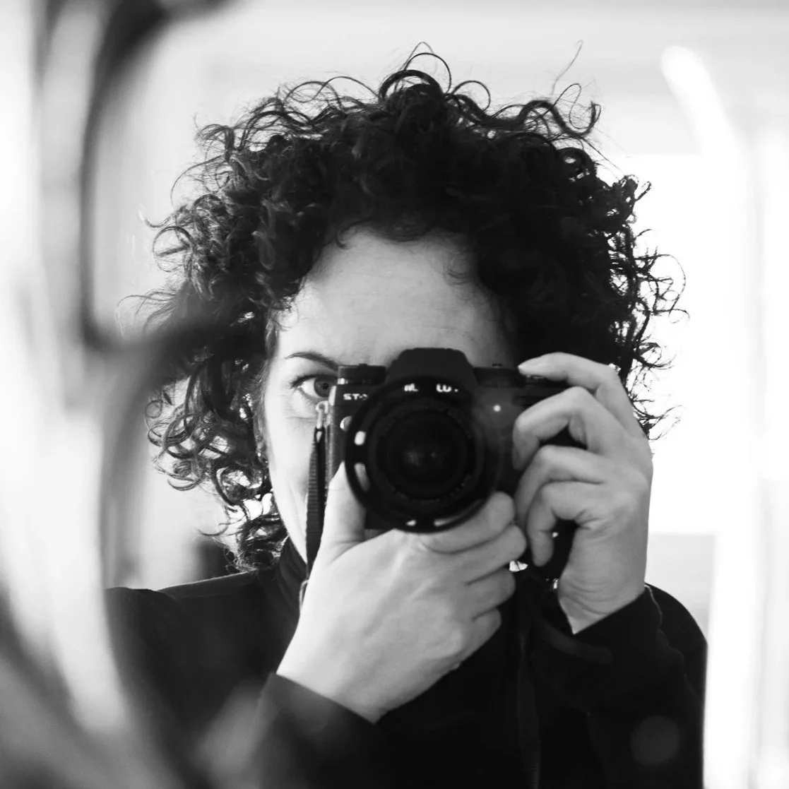 A woman with curly hair taking a photo with a camera reflected in a mirror, in black and white.