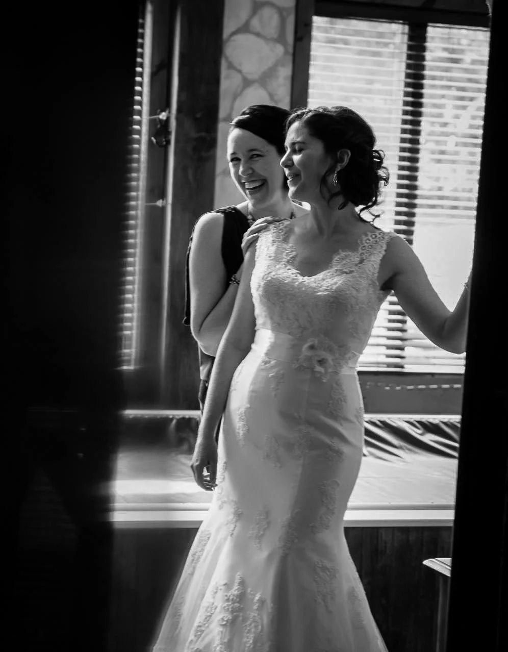 Two women, one in a wedding dress, smiling and sharing a joyful moment, standing near a window with blinds.