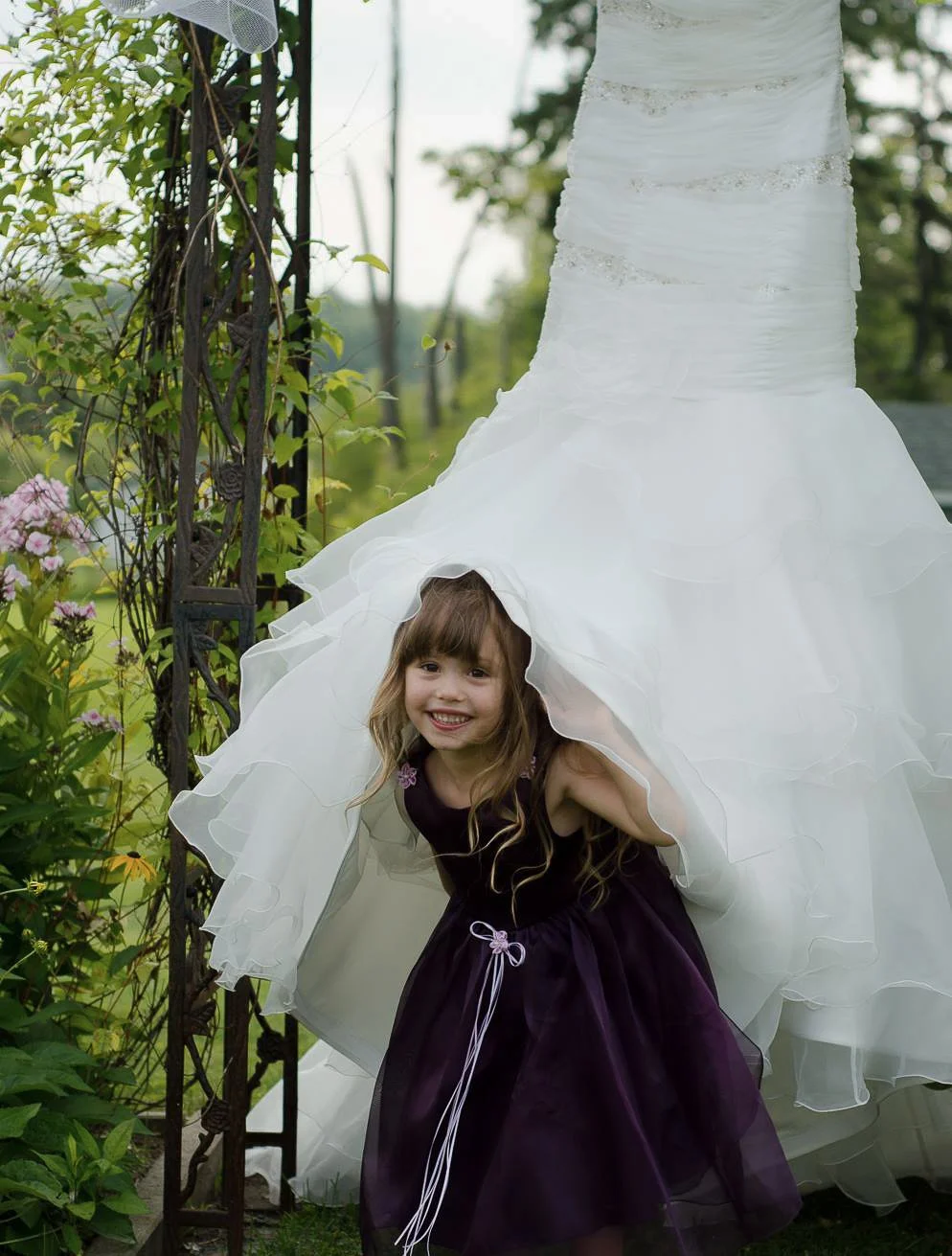 A young girl with long light brown hair, wearing a black dress with purple accents, is peeking out from under the hem of a white wedding dress hanging outdoors in a garden setting.