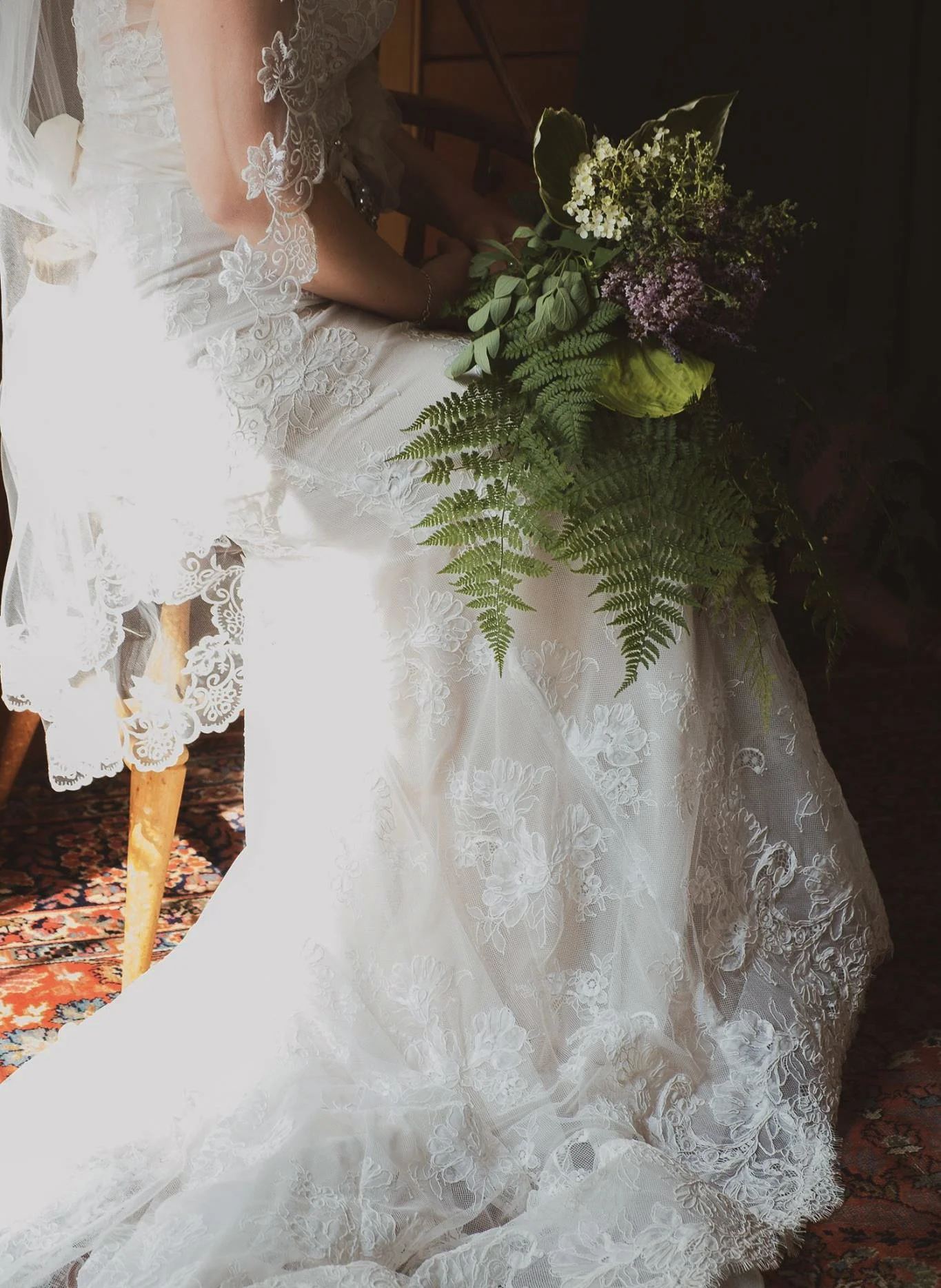A bride seated on a wooden chair, wearing a white lace wedding gown with floral embroidery, holding a bouquet of mixed flowers including white, purple, and green foliage. The gown features intricate lace details and a long train with lace accents. Th
